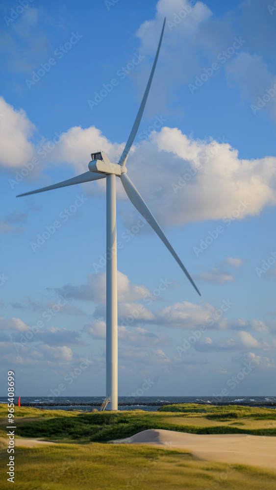 Wind turbine stands near the coast in the sand dunes, with its blades turning against a backdrop of blue skies and scattered clouds. Wind turbine stands near the coast in the sand dunes, with its blades turning against a backdrop of blue skies and scattered clouds.