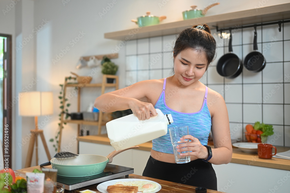 Beautiful young woman pouring milk into a glass while standing in a kitchen Beautiful young woman pouring milk into a glass while standing in a kitchen
