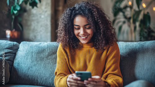 A happy young woman is relaxing on her couch at home, scrolling through her phone and smiling. She's using apps to play games, shop, and order food. photo