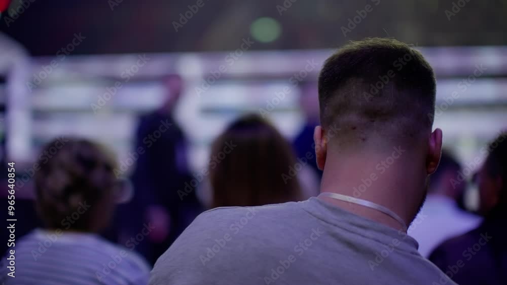Man watches a sporting event with friends in a dimly lit indoor stadium, illuminated by vibrant stage lighting. Excitement fills the air as the crowd cheers together Man watches a sporting event with friends in a dimly lit indoor stadium, illuminated by vibrant stage lighting. Excitement fills the air as the crowd cheers together