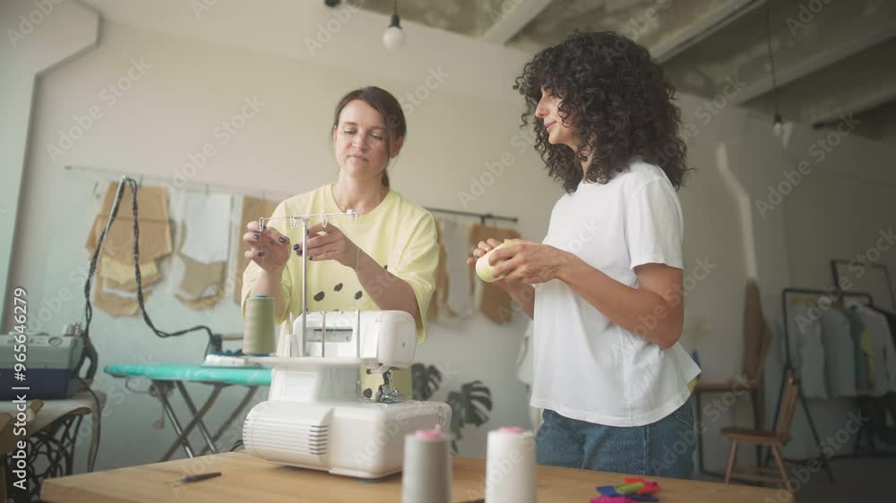 Hardworking seamstress teaching her assistant replacing threads in sewing machine. New employee holding spool of thread and watching mentor carefully. Working day in sewing factory. Small business. Hardworking seamstress teaching her assistant replacing threads in sewing machine. New employee holding spool of thread and watching mentor carefully. Working day in sewing factory. Small business.