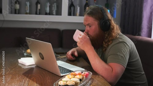 Millennial man sitting with a cup of coffee and a laptop in a city cafe with plate of rolls nearby watching an online video stream on the internet. photo