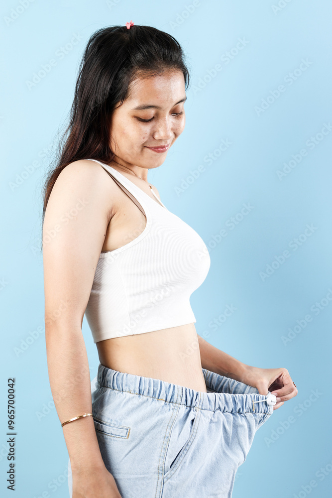 Indonesian Young Woman in Loose Clothes After Weight Loss on Blue Color Background Indonesian Young Woman in Loose Clothes After Weight Loss on Blue Color Background