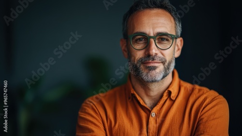 A confident man with greying hair and a beard, wearing green glasses and an orange shirt, stands indoors, epitomizing professionalism and maturity in a contemporary setting.