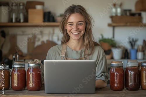 A happy woman sits at a wooden table with a laptop, surrounded by jars of homemade preserves in a rustic kitchen, exuding a warm, joyful ambiance.