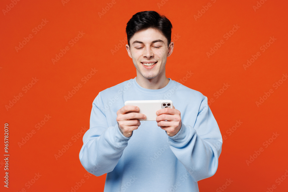 Young fun man wears blue sweatshirt casual clothes use play racing app on mobile cell phone hold gadget smartphone for pc video games isolated on plain red orange color background. Lifestyle concept. Young fun man wears blue sweatshirt casual clothes use play racing app on mobile cell phone hold gadget smartphone for pc video games isolated on plain red orange color background. Lifestyle concept.