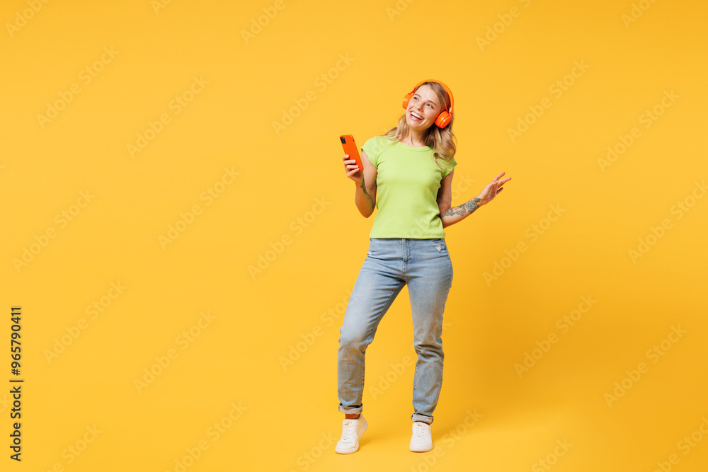 Full body young woman she wear green t-shirt casual clothes listen to music in headphones hold in hand use mobile cell phone look aside isolated on plain yellow orange background. Lifestyle concept. Full body young woman she wear green t-shirt casual clothes listen to music in headphones hold in hand use mobile cell phone look aside isolated on plain yellow orange background. Lifestyle concept.