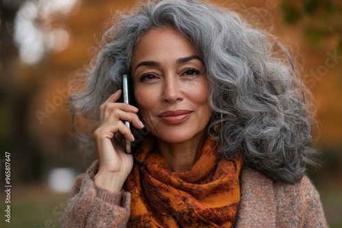 An older woman with beautiful curly gray hair, warmly dressed, smiles warmly while talking on the phone outdoors. Her joy and the autumnal background radiate warmth and connection. photo