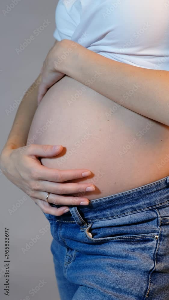 Video of young pregnant woman in white t-shirt and jeans on gray background. Belly exposed. Female on 5th month of pregnancy. Woman touching with hands her naked belly. Baby expectation concept. Video of young pregnant woman in white t-shirt and jeans on gray background. Belly exposed. Female on 5th month of pregnancy. Woman touching with hands her naked belly. Baby expectation concept.