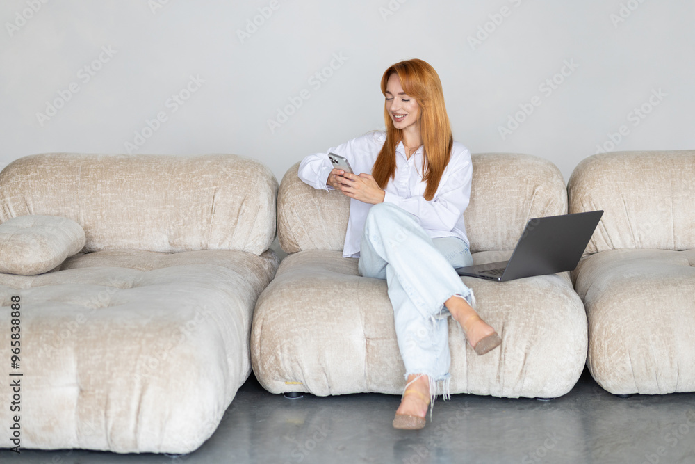 Young woman working with her laptop while using mobile phone sitting on a couch at home. Young woman working with her laptop while using mobile phone sitting on a couch at home.