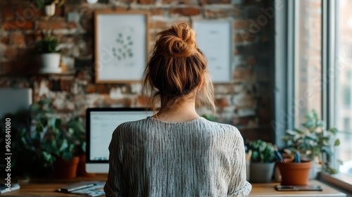 A person working diligently at their desk in a rustic home office with brick walls, embraced by plants and framed art, showcasing a cozy and productive workspace.