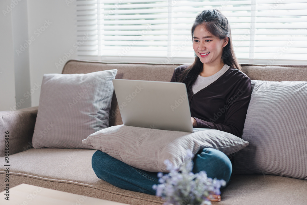 A young woman is sitting comfortably on sofa, using laptop while smiling. cozy atmosphere is enhanced by soft cushions and natural light streaming through window, creating warm and inviting space. A young woman is sitting comfortably on sofa, using laptop while smiling. cozy atmosphere is enhanced by soft cushions and natural light streaming through window, creating warm and inviting space.