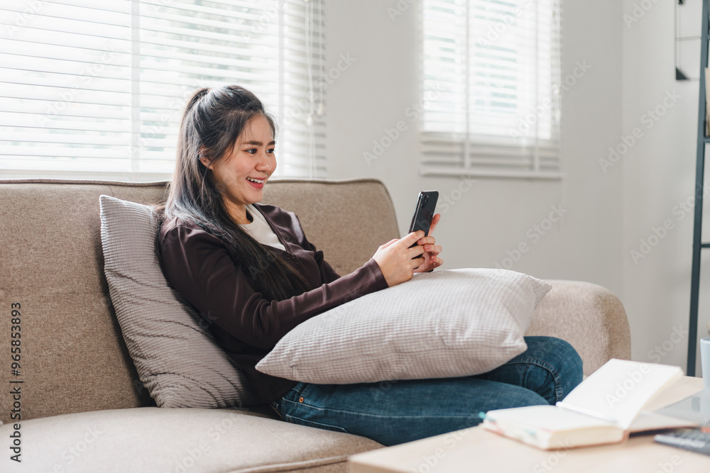 A young woman is sitting comfortably on sofa, smiling while using her smartphone. cozy atmosphere is enhanced by natural light streaming through window, creating relaxed vibe. A young woman is sitting comfortably on sofa, smiling while using her smartphone. cozy atmosphere is enhanced by natural light streaming through window, creating relaxed vibe.
