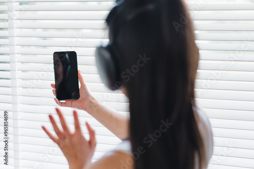 A woman wearing headphones is holding smartphone while standing by window with blinds, creating relaxed atmosphere. She appears to be engaged in virtual conversation, expressing emotions through her g photo