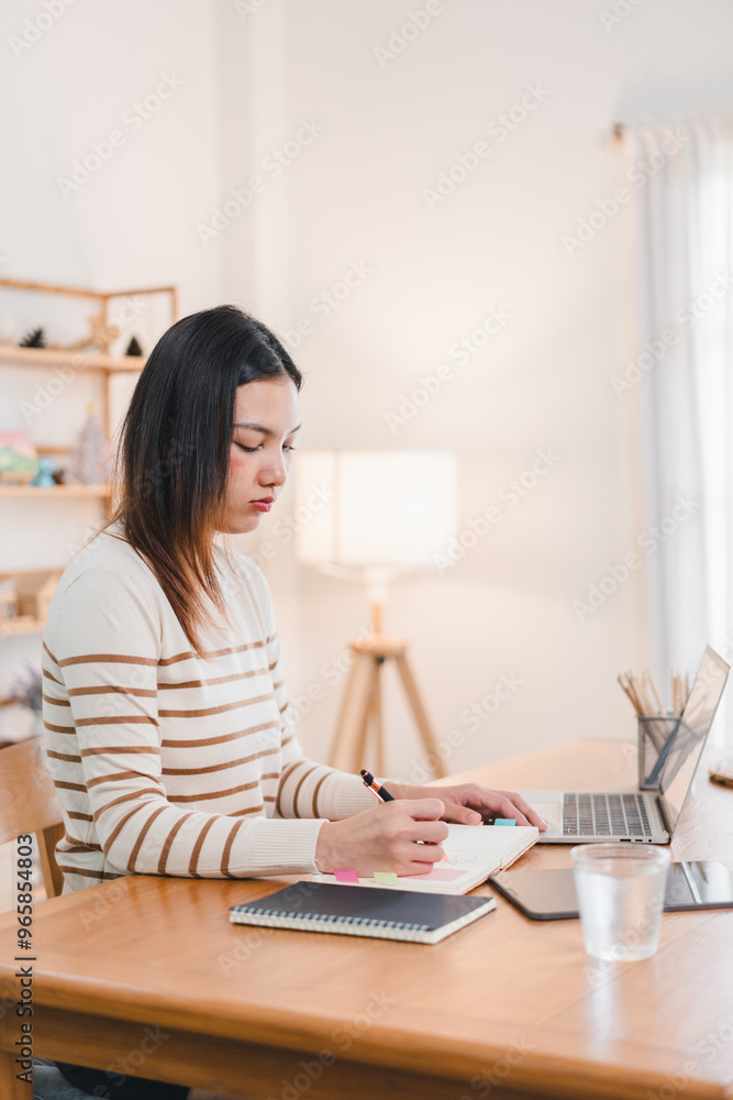 A woman is focused on her work at wooden desk, writing in notebook while using laptop. cozy atmosphere is enhanced by soft lighting and minimalist decor. A woman is focused on her work at wooden desk, writing in notebook while using laptop. cozy atmosphere is enhanced by soft lighting and minimalist decor.