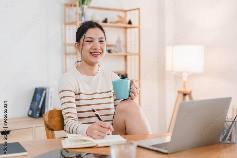 A young woman sitting at desk, smiling while holding blue mug and writing in notebook. cozy workspace features laptop and warm lighting, creating cheerful atmosphere. A young woman sitting at desk, smiling while holding blue mug and writing in notebook. cozy workspace features laptop and warm lighting, creating cheerful atmosphere.