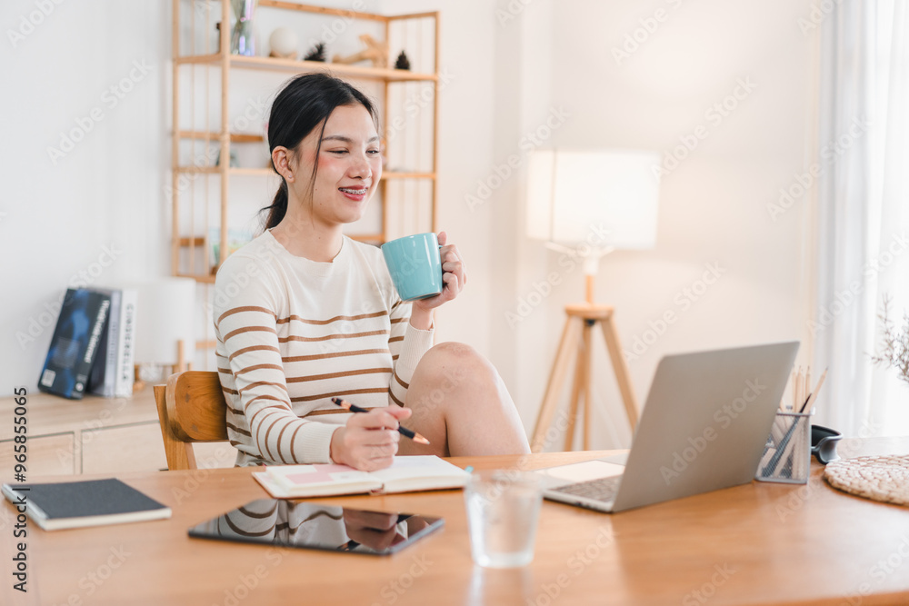 A woman sits at wooden desk, smiling while holding blue mug and taking notes. She enjoys cozy workspace with laptop and warm atmosphere. A woman sits at wooden desk, smiling while holding blue mug and taking notes. She enjoys cozy workspace with laptop and warm atmosphere.