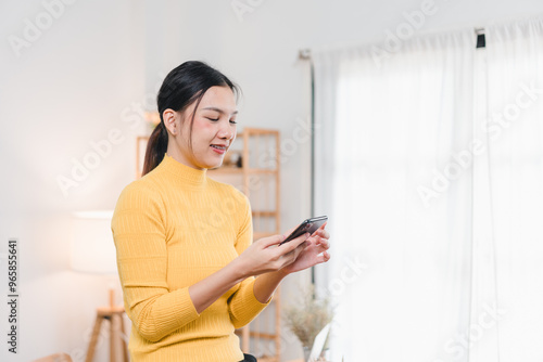 A young woman in yellow turtleneck smiles while using her smartphone in bright, cozy room. atmosphere is warm and inviting, perfect for connecting with friends or family.