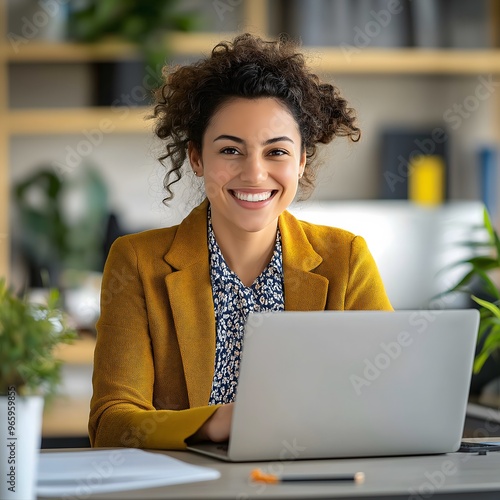 A stylish Middle Eastern manager with curly hair smiles warmly at the camera from her desk in a creative office, while colleagues go about their tasks in the background.