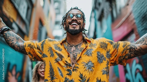 A smiling man with numerous tattoos raises his arms in joy while standing in a vibrant, graffiti-adorned street, embodying a spirit of freedom and exuberance, with a woman in background. photo