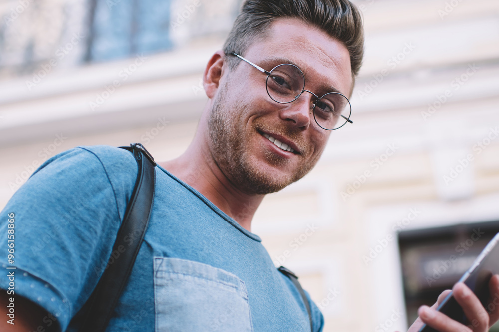 Below portrait of smiling male millennial with cell smartphone in hand enjoying free time for travelling, young man in spectacles for provide eyes protection using mobile phone for generation Below portrait of smiling male millennial with cell smartphone in hand enjoying free time for travelling, young man in spectacles for provide eyes protection using mobile phone for generation
