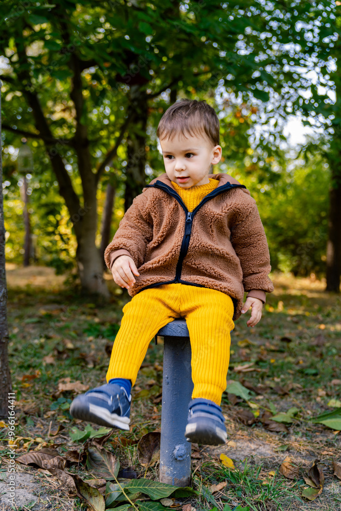 Toddler enjoying the outdoors in a forest setting during autumn. A young child sits on a small post, dressed warmly in cozy clothes