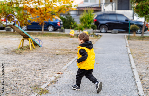 Young child in yellow vest walking through playground in early autumn. A child dressed in a yellow vest walks on a path in a playground surrounded by dry leaves photo