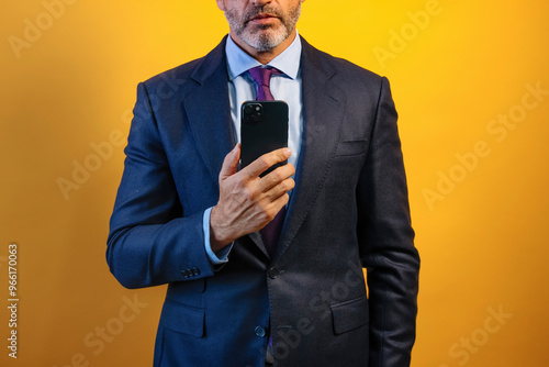 An unrecognizable man in a blue suit, using his smartphone, is standing in front of a yellow wall.