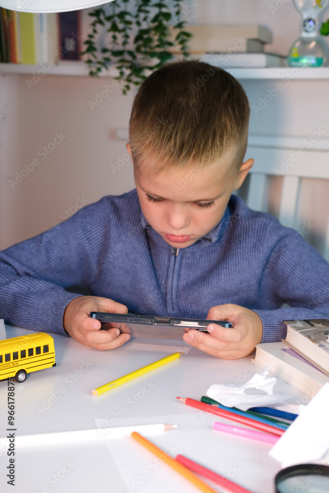 Child plays with smartphone instead of doing homework. Boy sits at desk and uses a mobile phone instead of reading a book. Education, school, learning disability, reading difficulties. Vertical image. Child plays with smartphone instead of doing homework. Boy sits at desk and uses a mobile phone instead of reading a book. Education, school, learning disability, reading difficulties. Vertical image.