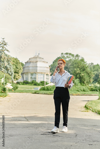 A teacher walks through a campus, talking on her phone and carrying books.