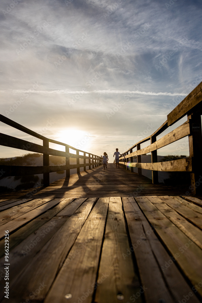 Naklejka premium Two children run along a wooden boardwalk bathed in the warm glow of the sunset. Capturing the essence of freedom, adventure and the beauty of nature.