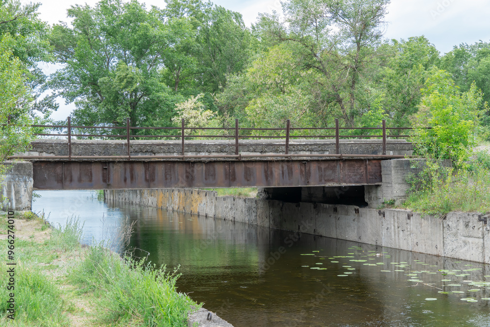Iron concrete bridge over the river canal. Reinforced pedestrian from one bank of water to other. Picturesque trees and bushes grow along the support slabs. Empty footbridge of bridge-canal with pier. Iron concrete bridge over the river canal. Reinforced pedestrian from one bank of water to other. Picturesque trees and bushes grow along the support slabs. Empty footbridge of bridge-canal with pier.