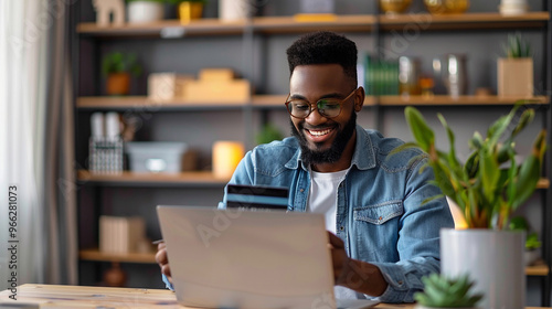 Man holding a credit card, using a laptop for online shopping in a modern home office.