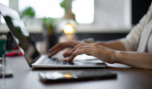 Close-up of female typing on a laptop in a well-lit workspace, with a smartphone resting on the desk. The background features plants and soft lighting, creating a productive and business atmosphere. photo