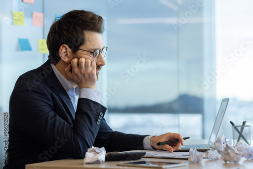 Businessman sitting at office desk appears bored while working on accounting tasks. He is using laptop and holding pen, surrounded by crumpled paper. Represents tedious office work and frustration. photo