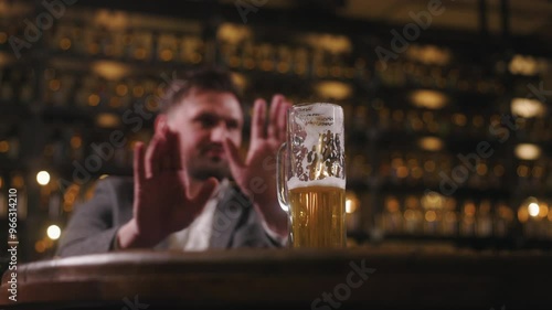 A man sitting in a bar or pub is refusing a glass of beer by raising his hand in a gesture of denial. The focus is on the half-full beer glass, with foam marks visible on the inside. photo