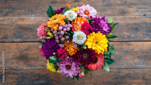 A top-down view of a vibrant bouquet of mixed flowers arranged in a round shape, with a variety of colors and textures, set on a wooden table
