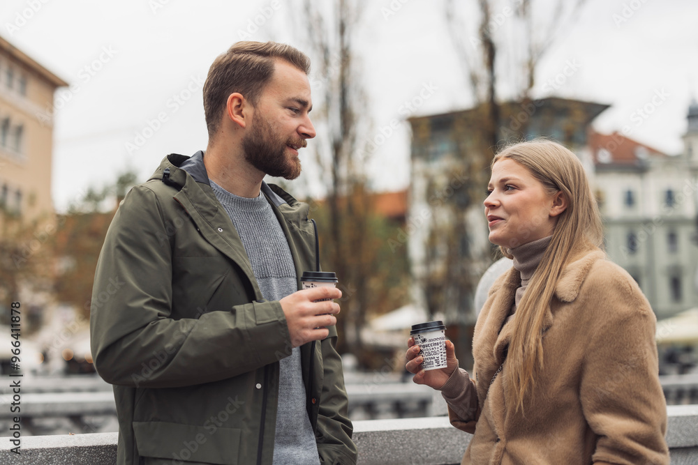Fototapeta premium A young couple enjoying coffee outdoors in an urban setting. They are engaged in conversation, with buildings and trees in the background, creating a cozy atmosphere.