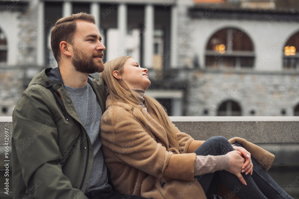 Obraz premium A couple sitting together outdoors, enjoying a moment of relaxation. The man has a beard and is wearing a green jacket, while the woman is dressed in a beige coat, leaning back against him. They are i