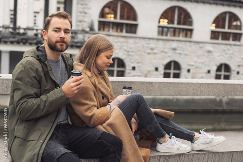 A young couple sitting on a ledge by a river, enjoying coffee. The man, wearing a green jacket, holds a coffee cup and looks at the camera. The woman, in a beige coat, is focused on her phone. The bac