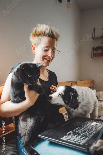 A person with short blonde hair and glasses sits on a couch, holding a black cat. A dog sniffs around a laptop on their lap. The setting is cozy with soft lighting.
