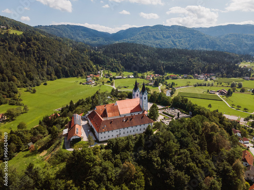 Aerial view of a historic monastery surrounded by lush green hills and valleys. The building features a red-tiled roof and two prominent towers. Nearby, small villages and winding roads are visible, s