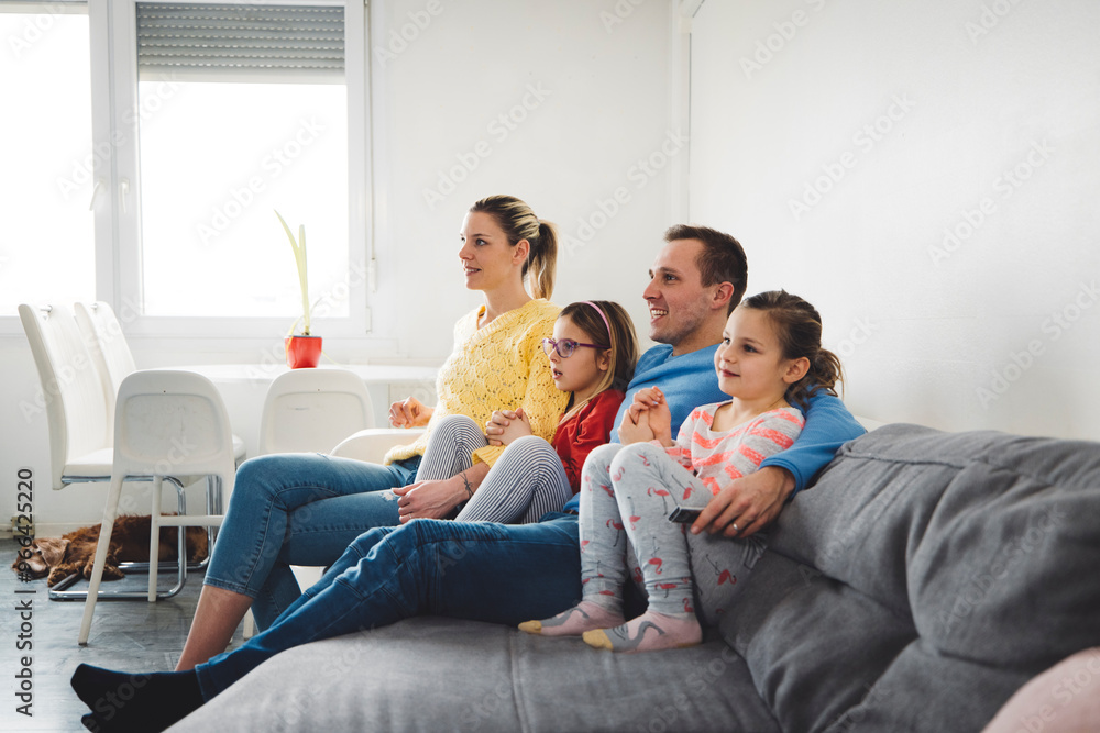 A family of four sitting together on a couch, watching TV. The mother and father are in casual clothing, with two young daughters beside them. The room has a bright, modern decor with a plant on the w A family of four sitting together on a couch, watching TV. The mother and father are in casual clothing, with two young daughters beside them. The room has a bright, modern decor with a plant on the w