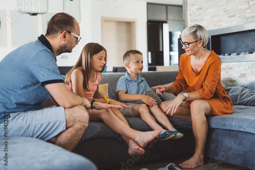 A family of four sitting on a modern gray sofa in a bright living room. They are engaged in a playful activity, with the parents and two children smiling and interacting. The atmosphere is warm and ch