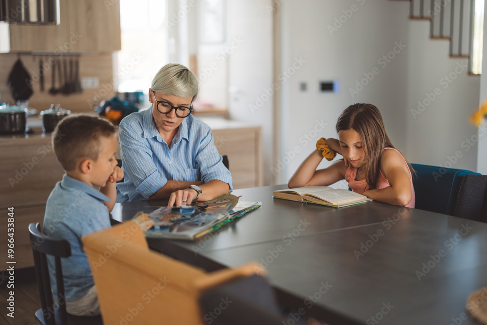 Fototapeta premium A woman mother with short blonde hair and glasses reads a book with two children at a dining table, doing homework with them. One child is looking at the book, while the other is reading a separate bo