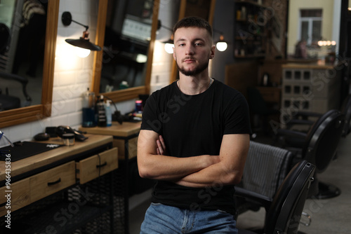 Confident young professional hairstylist with crossed hands at workplace, poses and looks at camera indoors barbershop. Male barber is ready to cut hair or beard. Men salon with mirrors, black chairs