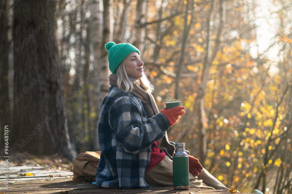 Pleased thoughtful woman rest alone walking in fall forest, drinking hot tea from thermos sitting on wooden path. Satisfied relaxed blonde female in warm clothes enjoys stroll in autumn woodland park Pleased thoughtful woman rest alone walking in fall forest, drinking hot tea from thermos sitting on wooden path. Satisfied relaxed blonde female in warm clothes enjoys stroll in autumn woodland park