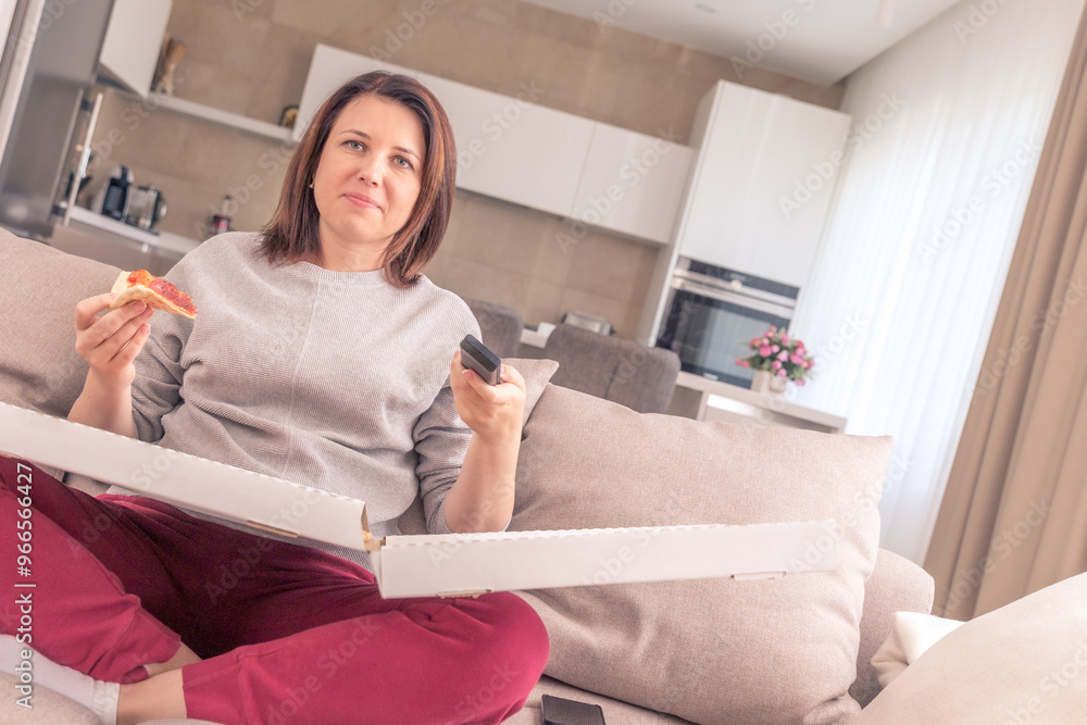 Beautiful woman eating pizza and watching TV holding remote control at home, orange tone Beautiful woman eating pizza and watching TV holding remote control at home, orange tone