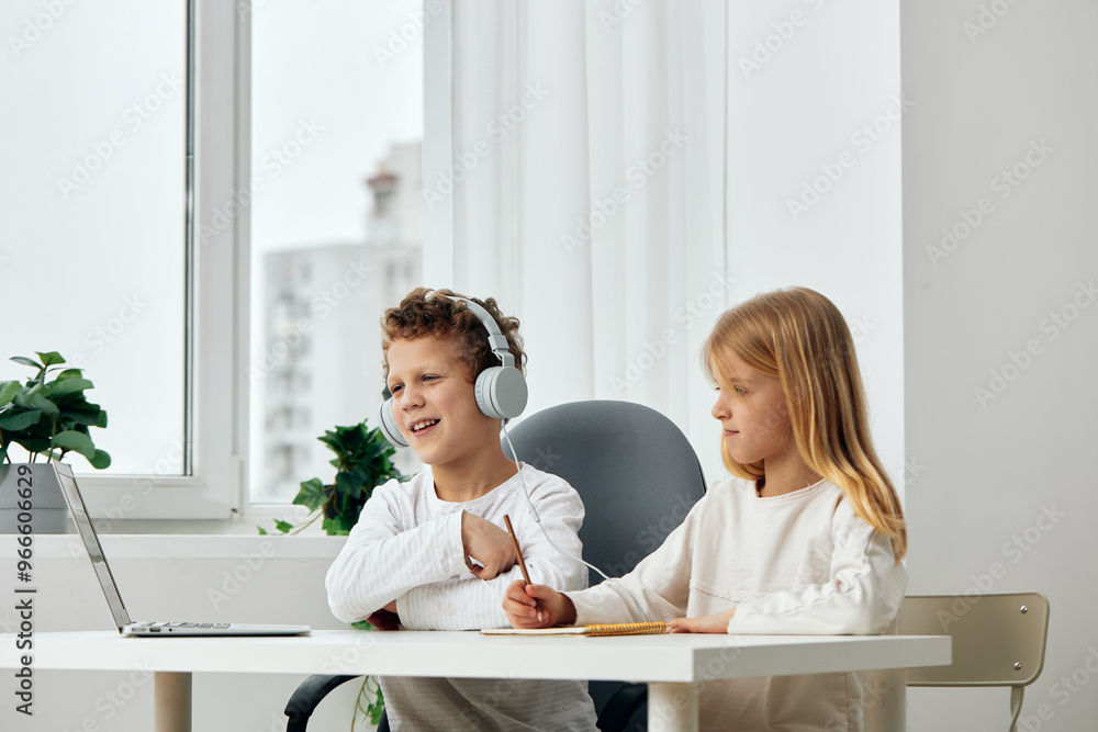 Happy Caucasian boy and girl studying and playing together at home They are sitting at a table in the living room, using their laptops and wearing headphones for an online elearning session The Happy Caucasian boy and girl studying and playing together at home They are sitting at a table in the living room, using their laptops and wearing headphones for an online elearning session The