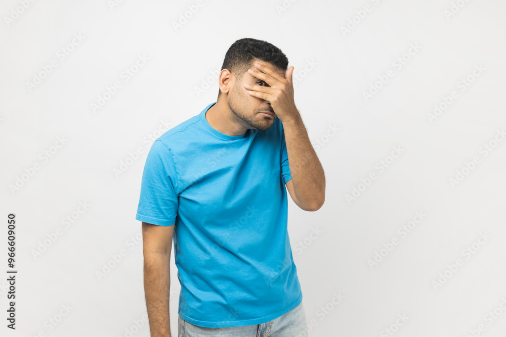 Portrait of curious attractive unshaven man wearing blue T- shirt standing looking at camera through fingers, peeking, sees something shameful. Indoor studio shot isolated on gray background. Portrait of curious attractive unshaven man wearing blue T- shirt standing looking at camera through fingers, peeking, sees something shameful. Indoor studio shot isolated on gray background.
